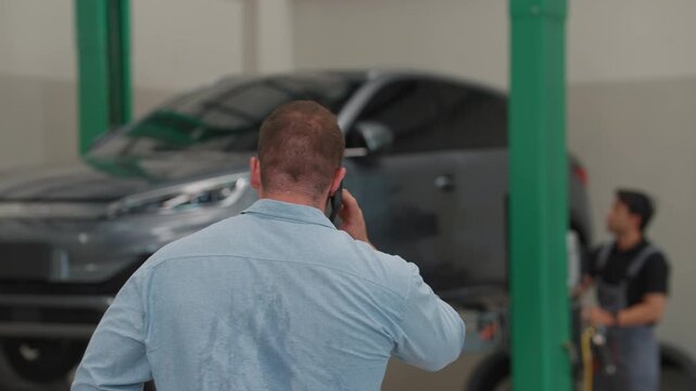 Man talks on phone near raised car. Worker stands nearby, observing the process. Scene shows garage operations in progress