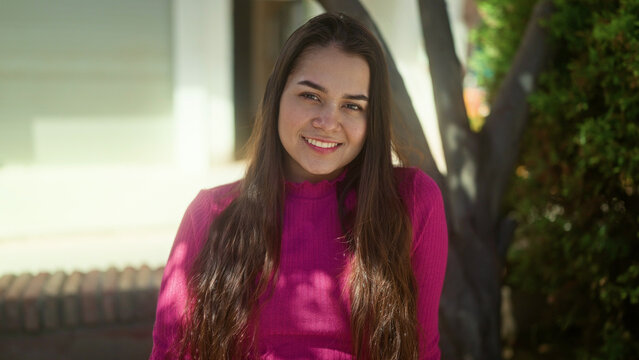 Woman smiling showing face and long hair in front of building wearing a pink sweater and natural sunlight; happiness comfort.