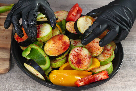 Hands in Black Gloves Mixing Sliced Raw Vegetables with Red Spices in a Round Pan