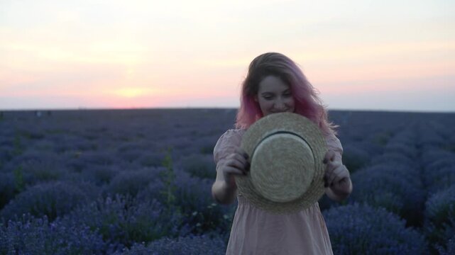 Woman playing with straw hat in lavender field at sunset, pink haired girl hiding face behind hat and smiling. Playful summer mood concept, pink sky over purple rows