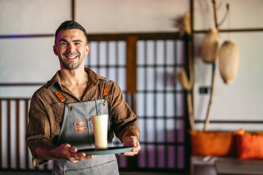 Young smiling handsome man waiter serving fruit juice, smoothie while working in a cafe.