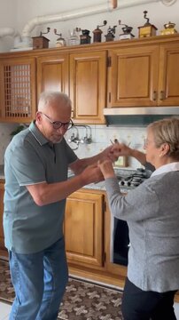 Joyful senior man and woman holding hands while dancing romantically in their home kitchen, celebrating their lasting love and companionship during retirement with smiles and gentle movements