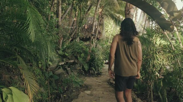 A barefoot man strolls along a sun-dappled stone path surrounded by dense tropical greenery and rustic thatched cabins, capturing peaceful retreat vibes in warm, natural daylight.