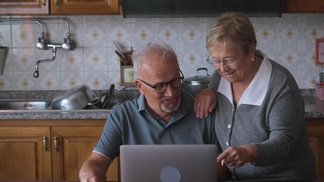 Happy elderly couple surfing the web on a laptop computer together in their kitchen, the woman pointing at the screen as they smile and discuss what they are watching online