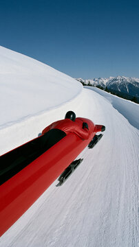 A bright red bobsled racing down a snow-covered track on a clear, sunny day with majestic mountains in the background