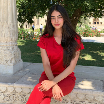 A young woman with long dark hair, wearing a red top and red pants, poses outdoors near a stone column with a garden in the background