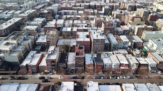 A sideways drone sweep crosses closely packed city roofs in Queens. Pale flat roofs form compact patterns between brick apartment blocks while parked cars and bare trees line narrow streets below.