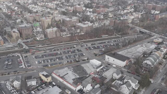 An aerial sweep follows railway platforms beside a full parking lot in Westchester County, New York. Parallel tracks border rows of cars with neighborhood buildings and bare trees in winter light.
