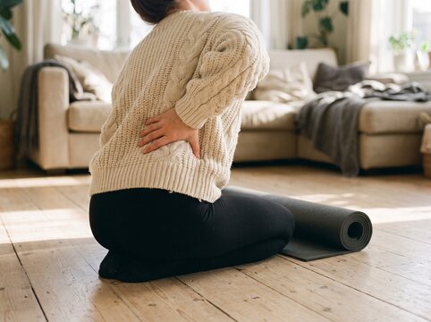 Woman suffering from lower back pain while sitting on a living room floor. Close-up shot of her hand holding her sore lumbar spine after a home workout. Lifestyle photography for health care.