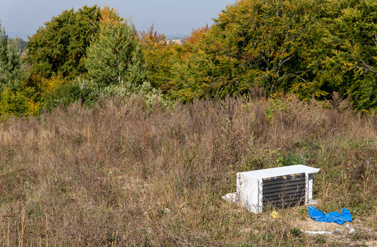 Old refrigerator with exposed cooling coils abandoned in overgrown grass near an autumn forest. Illegal appliance dumping, electronic waste and environmental pollution.