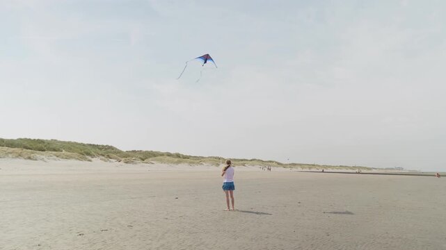 Girl flying a kite on a wide sandy beach near dunes slow motion rear view forward camera movement