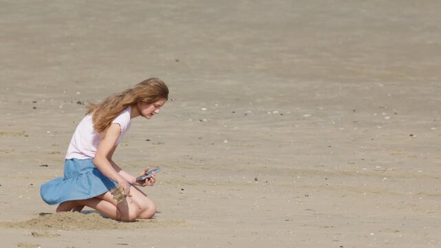 Girl digging sand and holding a smartphone by the sea