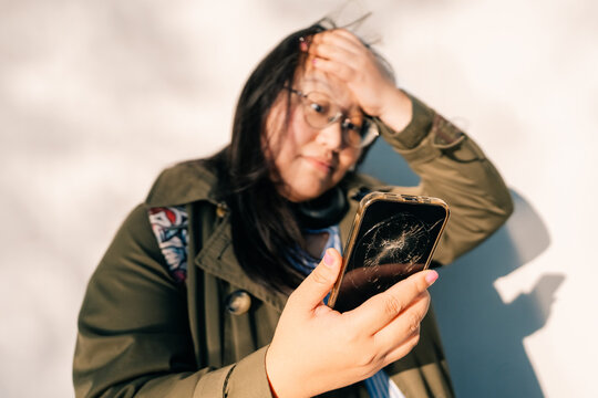 Woman looks at broken phone, standing against wall during daytime