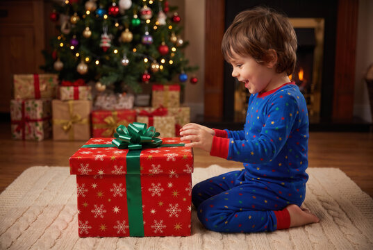 Smiling child in blue star pajamas reaches toward a closed red Christmas gift beside a decorated tree, capturing the eager anticipation of a magical surprise before the ribboned lid is opened