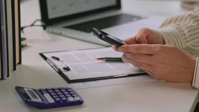 Person reviewing a printed contract while lifting a smartphone beside a calculator and laptop on an office desk during document preparation. contract preparation process