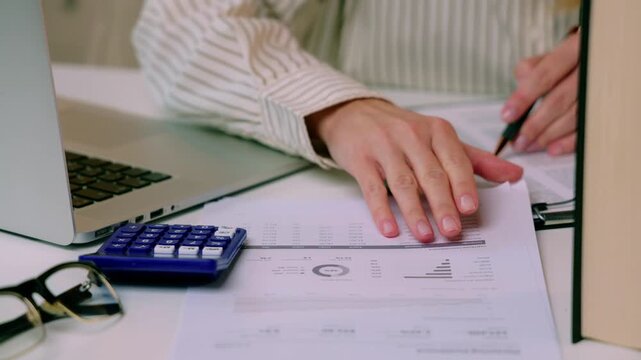 Person reviewing financial documents and charts while turning pages beside laptop and calculator at office desk. checking financial figures for accounting analysis and business report preparation in o