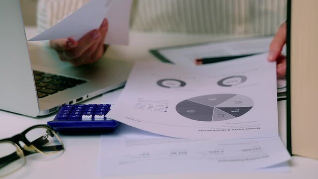 Person reviewing printed financial charts and moving reports beside laptop and calculator during desk based analysis. checking financial figures for accounting analysis and business report preparation