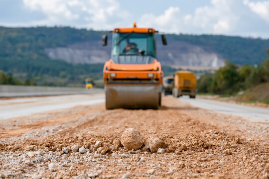 Orange roller moves across freshly laid road. Driver operates machine with focused attention. Scattered gravel lies in foreground. Mountain range forms distant backdrop