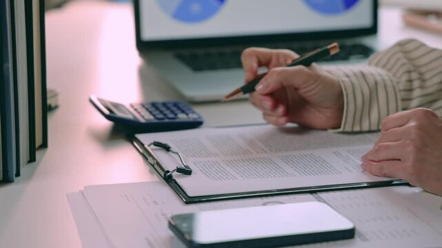 Person reviewing and signing contract documents with pen at office desk with laptop, calculator, and smartphone. showing contract signature preparation and approval workflow