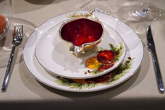 Festive table setting with ceramic plates decorated with pomegranates, orange slices and pine branches, golden pomegranate bowl in center. Christmas and New Year table decor in red and gold colors.