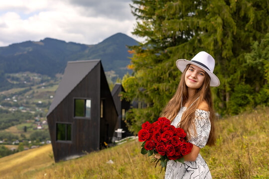Beautiful young woman in white hat holding a bouquet of red roses in scenic mountain countryside, romantic outdoor portrait near modern cabins surrounded by green forest.