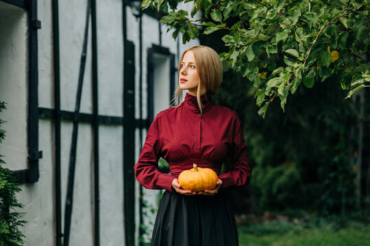 Woman in vintage dress holds a pumpkin outdoors by an old house
