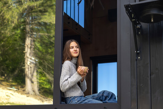 Young woman in gray sweater sitting by an open window holding a cup and enjoying mountain scenery, peaceful morning relaxation in a modern cabin surrounded by forest.