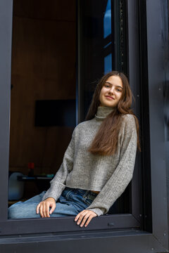 Beautiful young brunette woman in gray knitted sweater and blue jeans sitting in an open window, relaxed female portrait with a gentle smile at a modern dark cabin.