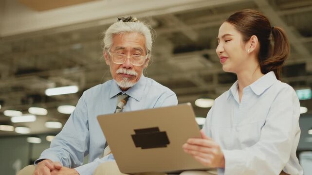 Young Thai woman brings laptop closer to senior man and both smile