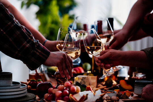 Toast with glasses of white wine during festive lunch meal. Gourmet food pairing with artisan cheese board at dinner table. Friends gathering, party and hospitality concept.