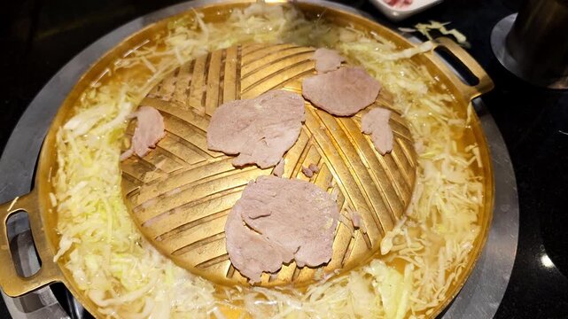 Chopsticks lift a thin slice of meat from a traditional brass Thai barbecue pan, or mu kratha, featuring a central dome for grilling and a trough of simmering broth with cabbage.