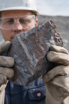 Male miner holding a raw metal ore sample in protective gloves and safety equipment. Mining industry, mineral extraction, geology inspection and industrial resource concept.