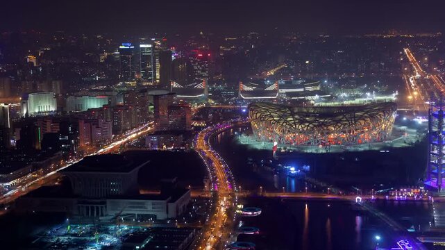 Aerial night view of Beijing&rsquo;s Olympic Park featuring the illuminated Bird&rsquo;s Nest stadium, surrounding high-rises, and traffic trails along city highways