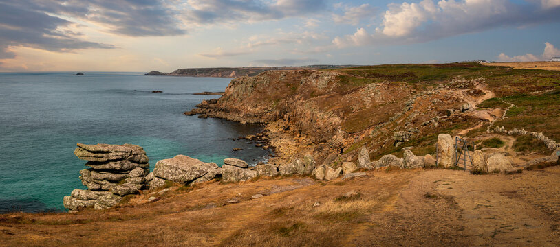 Panoramic view of two people sitting on a granite outcrop above a turquoise cove on the Cornish coast path near Land&rsquo;s End, Cornwall, England, in warm evening light