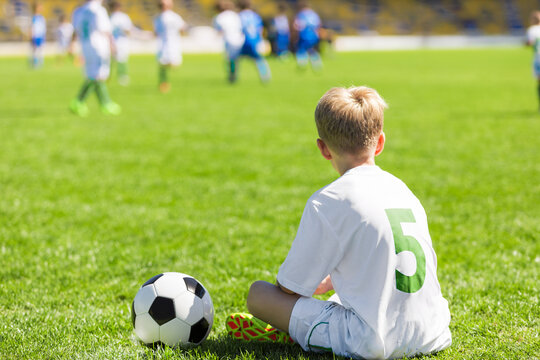 Player Number 5 Sitting on Grass Next to Soccer Ball. Young Soccer Substitute Player in White Jersey Number 5 Sitting on Grass Pitch Next to Ball Watching Match on Field