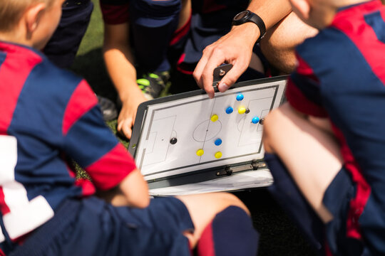 Soccer Coach Explaining Tactics on Board to Team in Blue/Red Kit. Youth Soccer Coach Using Tactical Clipboard to Explain Game Strategy to Junior Players in Blue and Red Uniforms During Match Break