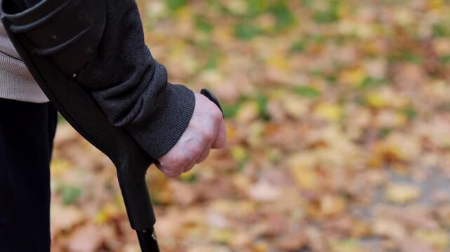 Close-up of an elderly man's hand gripping a forearm crutch. Senior patient in rehabilitation walking outdoors in an autumn park after hospital discharge.