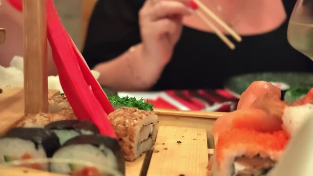 Close-up dining scene showing hand reaching for shot glass of dark soy or dipping sauce on wooden sushi platter surrounded by assorted sushi rolls, salmon sashimi, bright orange fish roe, imitation kr