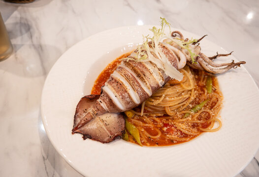 Grilled whole squid spaghetti pasta with tomato sauce on white plate.