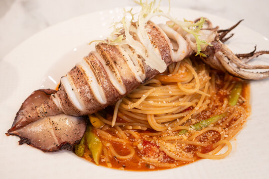Grilled whole squid spaghetti pasta with tomato sauce on white plate.