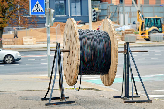 Wooden cable reel with wound black electrical wire mounted on metal support stands at an urban construction site. Spool of industrial cable on wooden drum metal supports, construction infrastructure