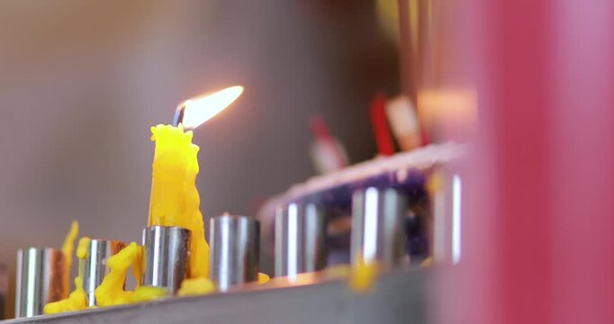 Burning yellow candle with melted wax and glowing flame, incense sticks and burner in the blurred background, symbolizing Buddhist worship, faith and religious rituals, close-up