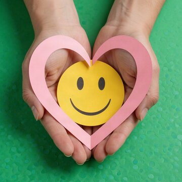 Hands holding a pink paper heart and yellow smiley face on green background