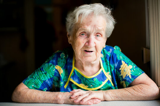 Portrait of an elderly woman resting her folded hands on a table by a window and looking at the camera indoors