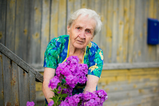 Portrait of an elderly woman leaning beside purple flowers outside a rustic wooden house and looking at the camera
