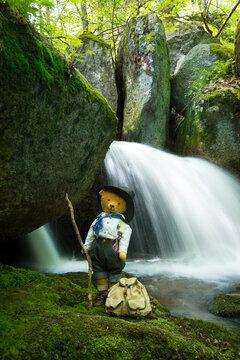 Vintage teddy bear dressed as traditional hiker with backpack and walking stick in front of forest waterfall