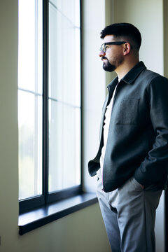 Man in casual attire standing by window, looking outside. Reflecting, thinking, planning future, enjoying view