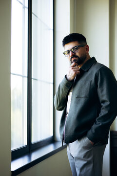 Young man wearing a jacket and glasses, thinking deeply by a large window, conveying concepts of contemplation and focus