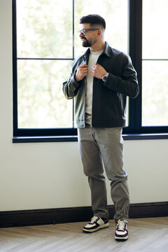 Man in casual fashion style standing by a large window, wearing a jacket, t-shirt, and pants, presenting a modern look