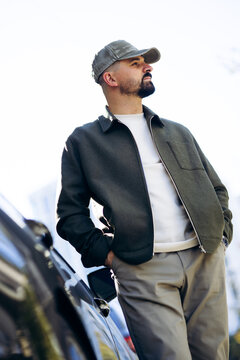 Man wearing a jacket and cap, leaning on a car, looking away, with hands in pockets. Reflecting modern lifestyle and confidence outdoors
